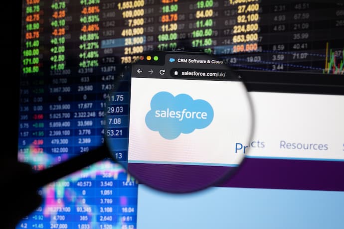 Salesforce company logo on a website with blurry stock market developments in the background, seen on a computer screen through a magnifying glass.